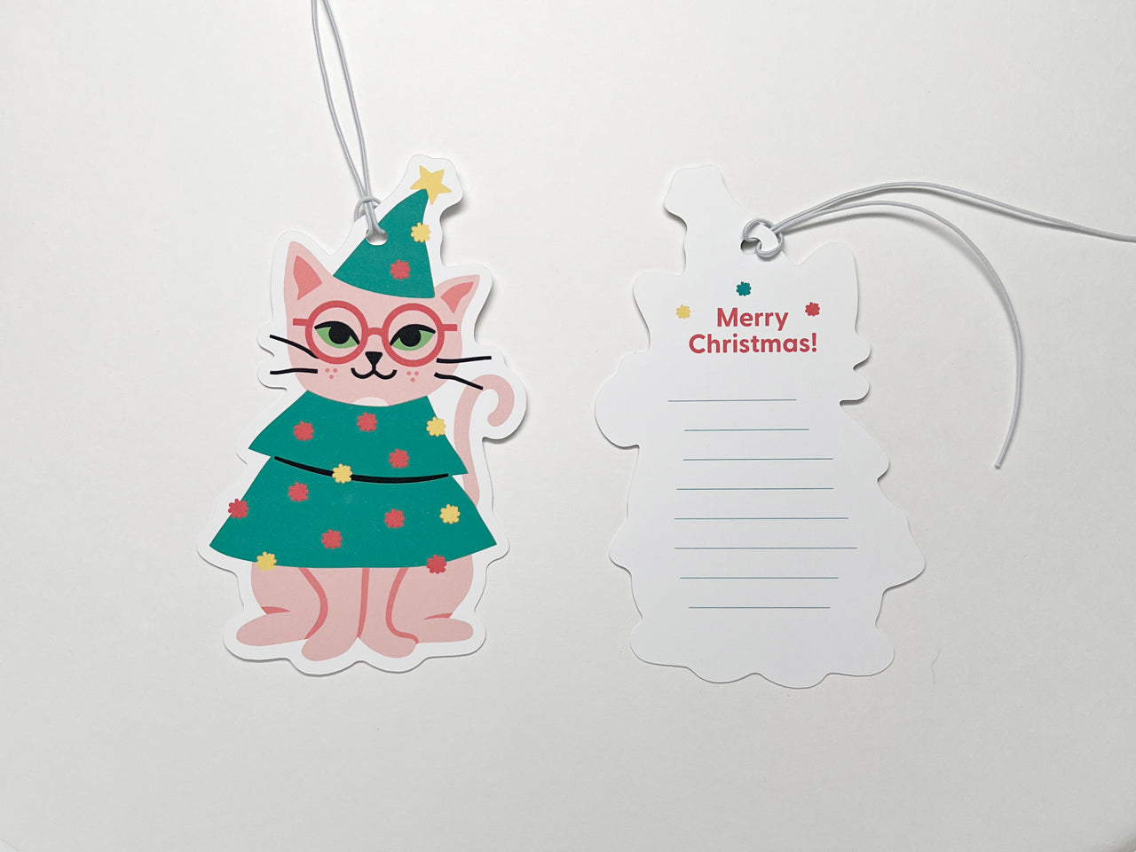Two holiday gift tags featuring a cute cat dressed as a Christmas tree. The cat, with pink fur and glasses, wears a green tree outfit with colorful ornaments and a star on top. One tag displays the cat illustration, while the back of the other tag shows "Merry Christmas!" with space for a personal message. Both tags have white strings, set against a plain white background that highlights the festive colors.