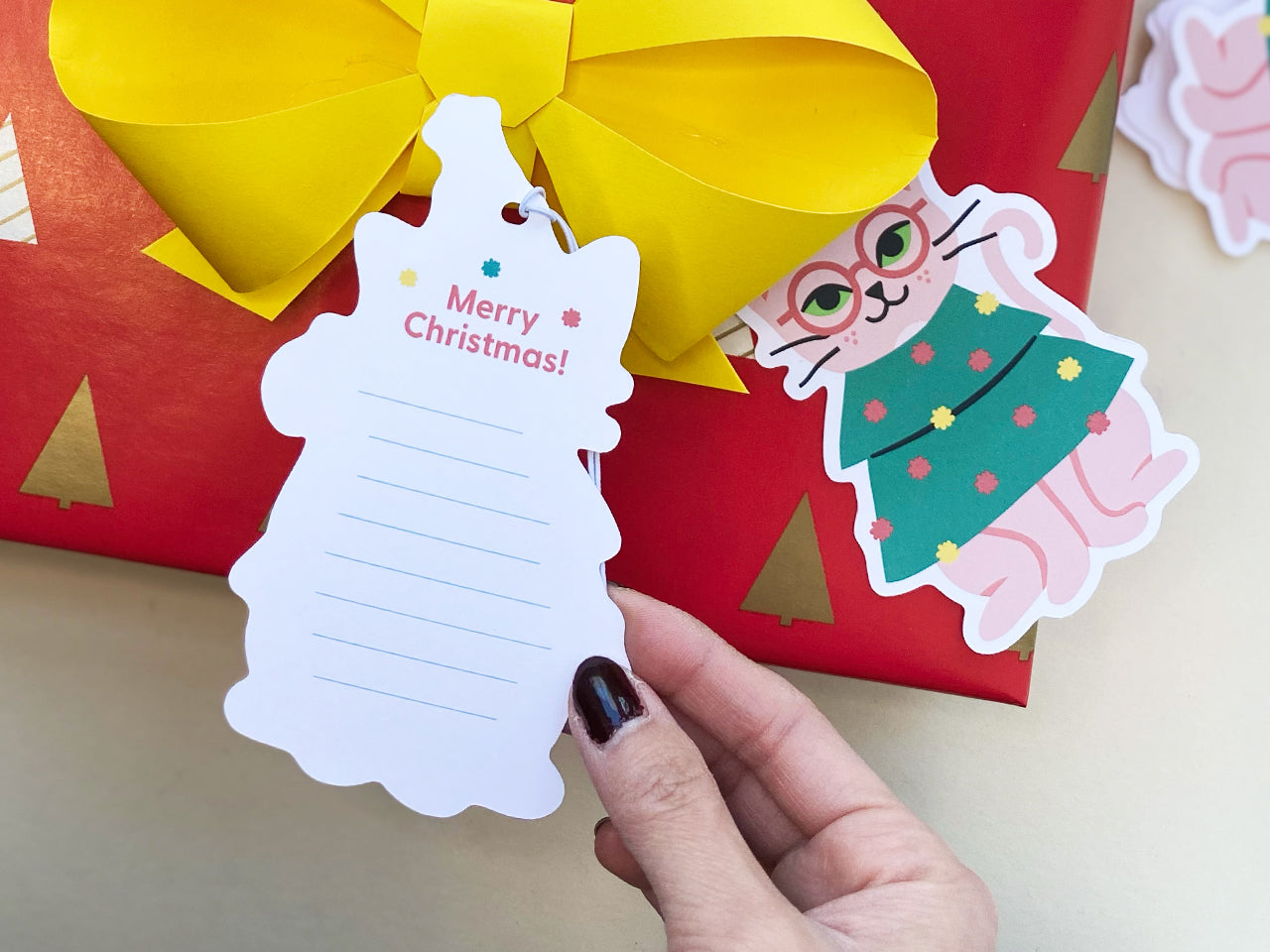 Close-up of a Christmas gift tag shaped like a festive tree, featuring the text 'Merry Christmas!' with space for writing. Paired with a cute illustration of a pink cat wearing glasses and a green Christmas tree outfit. Perfect for holiday gift wrapping.