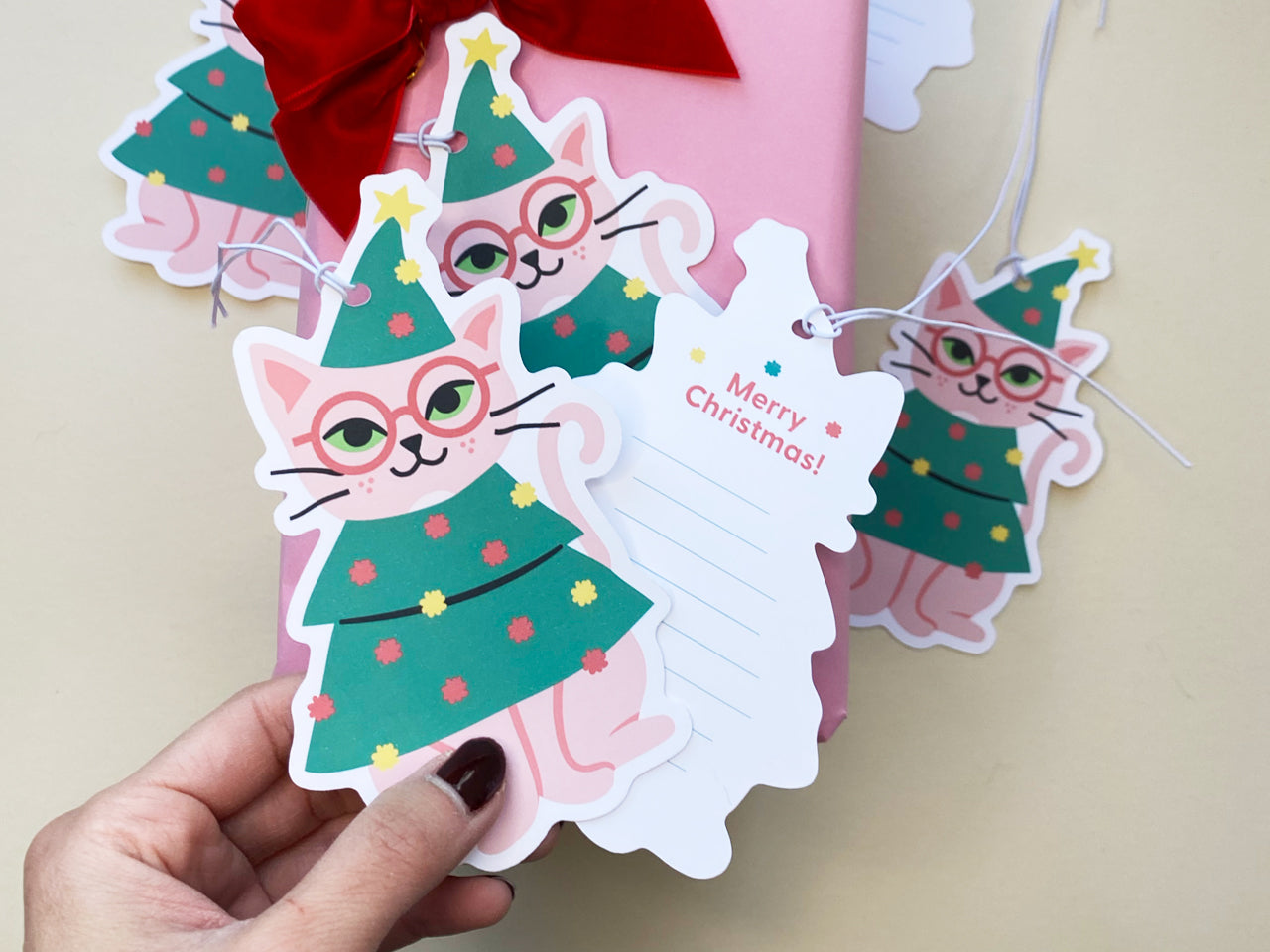 Festive Christmas gift tags featuring a playful pink cat dressed in a tree outfit with colorful decorations and glasses. Paired with matching writable 'Merry Christmas!' tags, perfect for wrapping gifts in style.