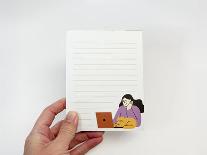 A hand holding a small lined notepad against a white background, featuring a minimalist illustration of a woman working on a laptop with an orange cat resting beside her, representing a work-from-home cat themed notepad.