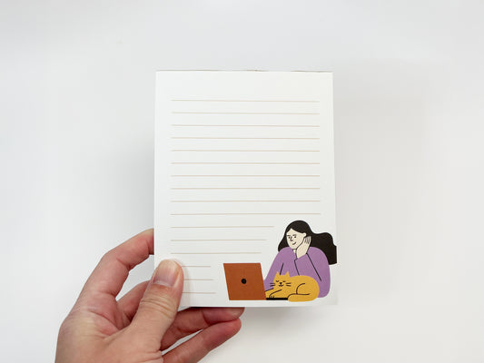 A hand holding a small lined notepad against a white background, featuring a minimalist illustration of a woman working on a laptop with an orange cat resting beside her, representing a work-from-home cat themed notepad.