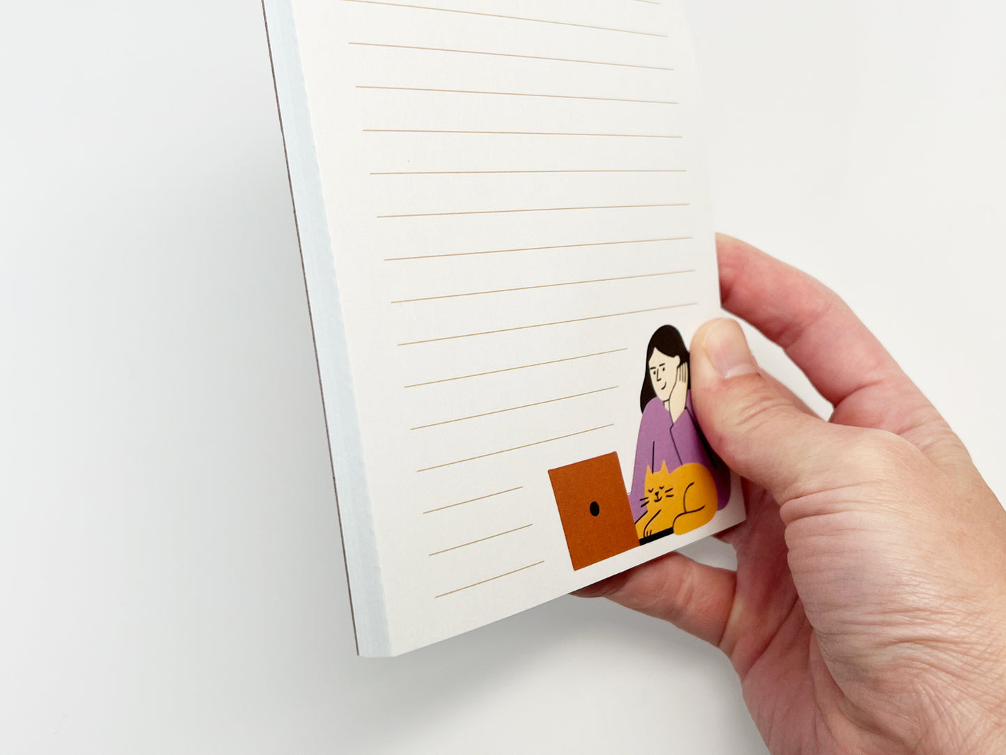 A close-up side view of a small lined notepad held in one hand, showing the thickness of the paper and a minimalist illustration of a woman working on a laptop with an orange cat resting beside it at the bottom corner.