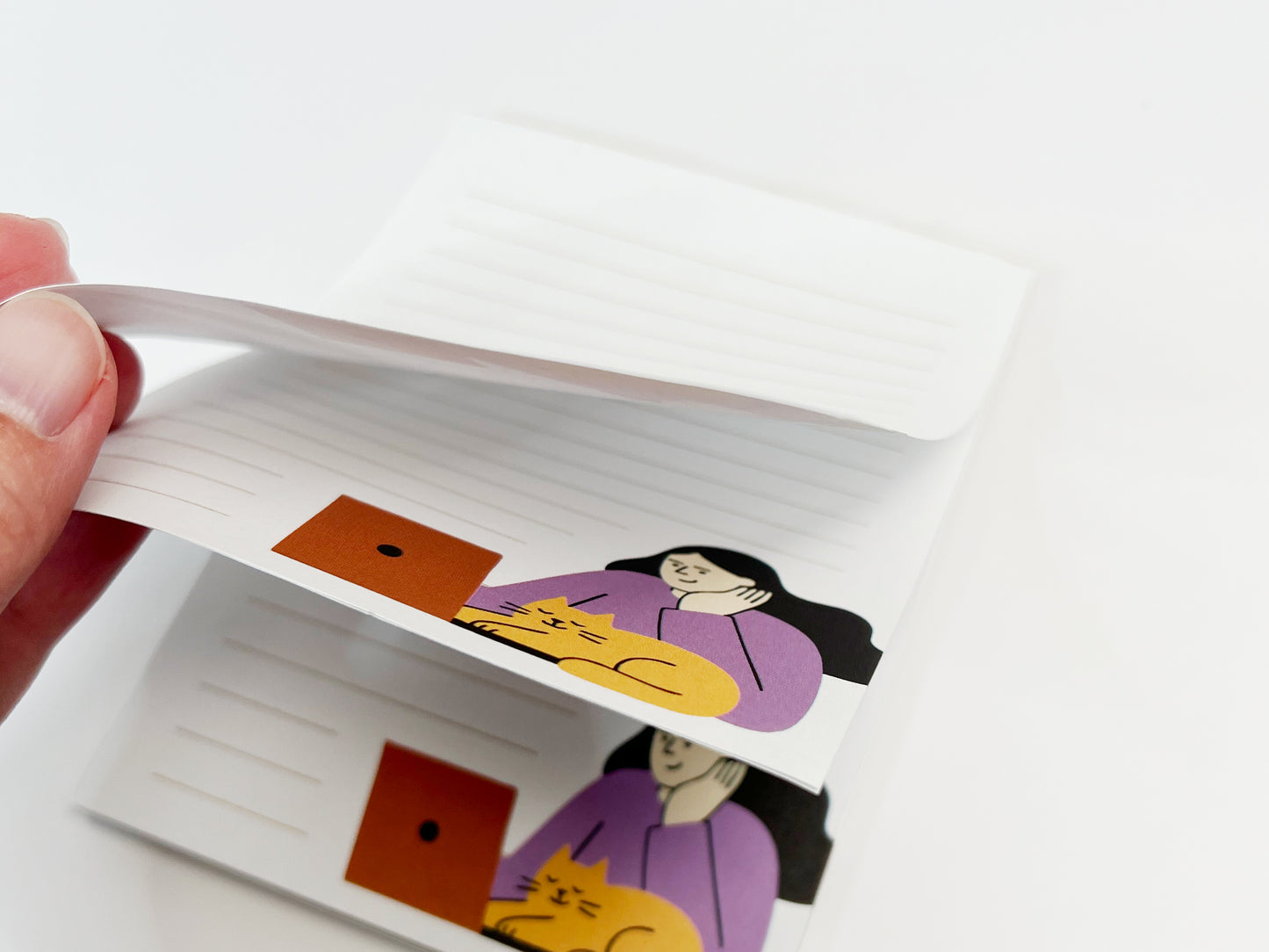 A close-up of a small lined notepad with pages slightly lifted, showing multiple sheets and a minimalist illustration of a woman working on a laptop with an orange cat resting beside her.