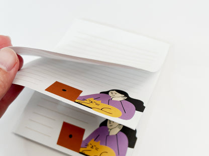A close-up of a small lined notepad with pages slightly lifted, showing multiple sheets and a minimalist illustration of a woman working on a laptop with an orange cat resting beside her.