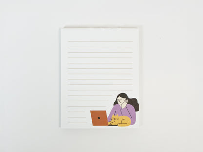 A small lined notepad on a white background featuring a minimalist illustration of a woman working on a laptop with an orange cat resting beside her, designed as a cute work-from-home cat notepad.