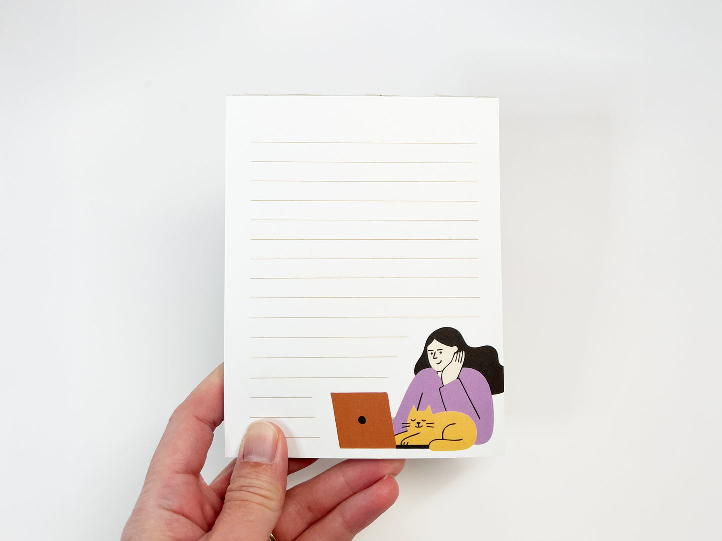 A hand holding a small lined notepad against a white background, featuring a minimalist illustration of a woman working on a laptop with an orange cat resting beside her, representing a work-from-home cat themed notepad.