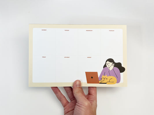 A horizontal 8.5 x 5.5 inch weekly planner notepad held in one hand against a white background, featuring an undated Monday–Sunday layout with clean sections, a soft beige border, and a playful illustration in the bottom right of a woman working on a laptop while a yellow cat lounges beside her, suggesting a cozy work-from-home setting.