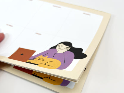 A close-up photo of a stacked weekly planner notepad showing multiple tear-off sheets, with an undated Monday–Sunday layout, a soft beige border, and a playful illustration of a woman working on a laptop while a yellow cat naps beside her, highlighting the paper thickness and layered pages against a white background.