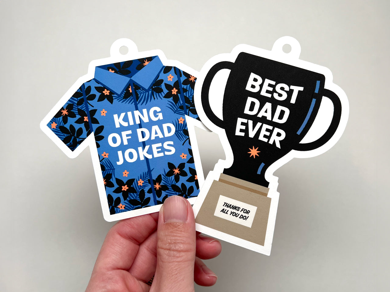 Hand holding up a "King of Dad Jokes" Hawaii Shirt illustration and "Best Dad Ever" trophy illustration as Father's Day gift tags.