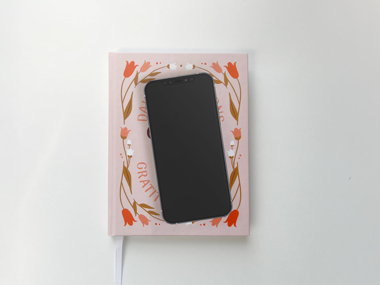 Personalized pink gratitude hardcover journal with a floral design placed beneath a black smartphone to show size. The front cover features an elegant floral border with red, white, and brown flowers surrounding customizable text for a name. The compact size is highlighted by the placement of the smartphone, suggesting easy portability and daily use. A white ribbon bookmark extends from the bottom, making it ideal for marking pages during journaling, mindfulness practices, or daily reflections.