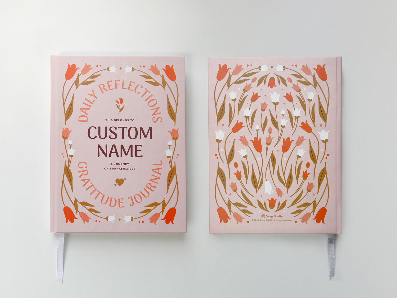 Two personalized gratitude journals with custom name options, displayed on a white background. The journal on top has a soft light pink cover featuring an elegant modern floral design. The text on the cover reads, Daily Reflections Gratitude Journal – A Journey of Thankfulness, with space to add a custom name in the center. The journal underneath has a calming purple cover adorned with a matching floral design. Both journals are hardcover and designed for mindfulness and daily reflections.