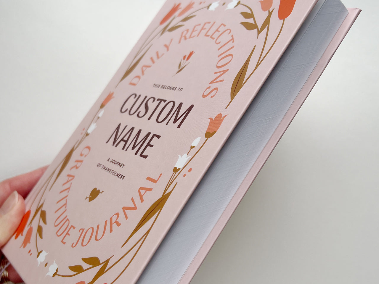 Close-up of a personalized pink gratitude journal being held at an angle, highlighting the sturdy hardcover construction and smooth, high-quality finish. The cover design includes delicate floral elements in red, white, and brown tones, surrounding customizable text. The edges of the pages show the thickness of the journal, emphasizing its durability and suitability for daily reflections and gratitude practice. The image conveys the craftsmanship that make the journal an ideal self-care gift.