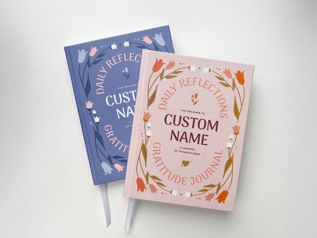 Two personalized gratitude journals with custom name options, featuring modern floral designs on hardcover. One journal has a light pink cover while the other has a purple cover with floral elements. Both journals include a white ribbon bookmark and are designed for daily reflections, mindfulness, and self-care. Ideal as thoughtful gifts for women, these customizable journals are perfect for journaling, affirmations, and gratitude practice. Styled on minimal white background for elegant presentation.