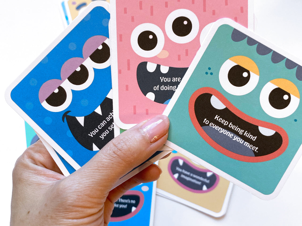 A hand holding three monster-themed lunch box note cards, each featuring a cheerful monster face and positive messages. More colorful cards are spread out in the background on a white surface.