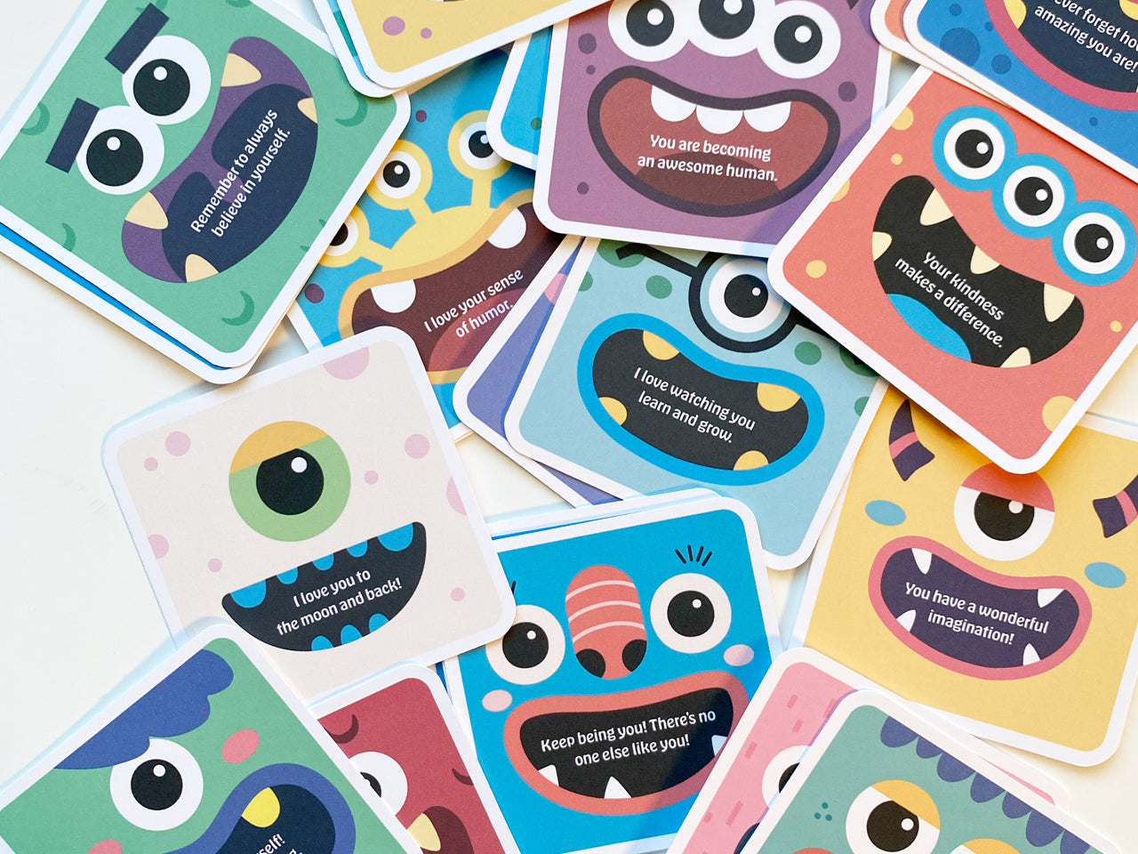 A flat lay of several colorful monster-themed lunch box note cards scattered across a white surface. Each card is square with round corners and features a unique cartoon monster design with bright colors and encouraging messages.