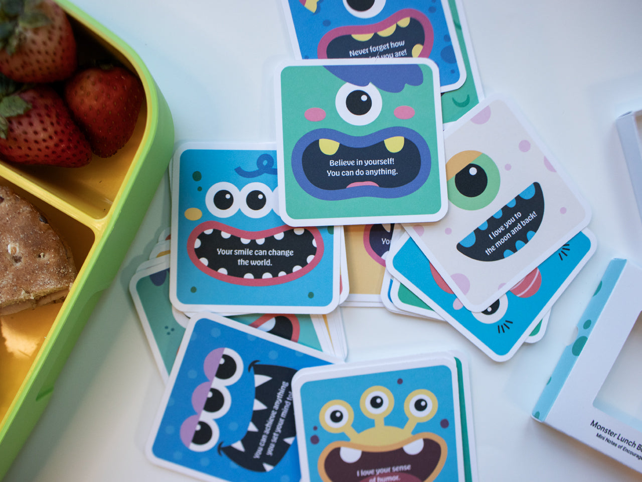 A variety of colorful monster-themed lunch box note cards scattered next to a green lunch box filled with strawberries and a sandwich. Each card features a whimsical monster and uplifting messages to motivate and inspire kids during school lunch.