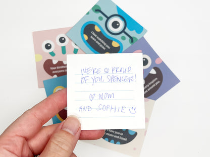 Hand holding a small square lunch box note with lined back and handwritten message, with colorful monster lunch notes blurred in the background, showing handmade kids lunchbox cards designed for school lunches and backpacks.