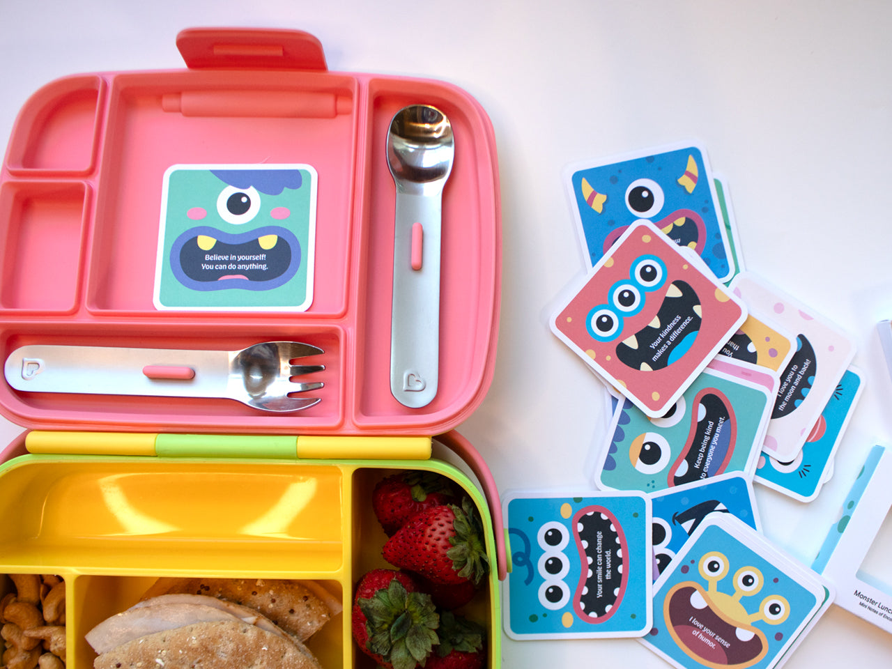 A pink and yellow lunchbox with a green monster note card placed inside, reading Believe in yourself! You can do anything. Surrounding the lunchbox are multiple colorful monster lunch box note cards with cheerful designs and encouraging messages.