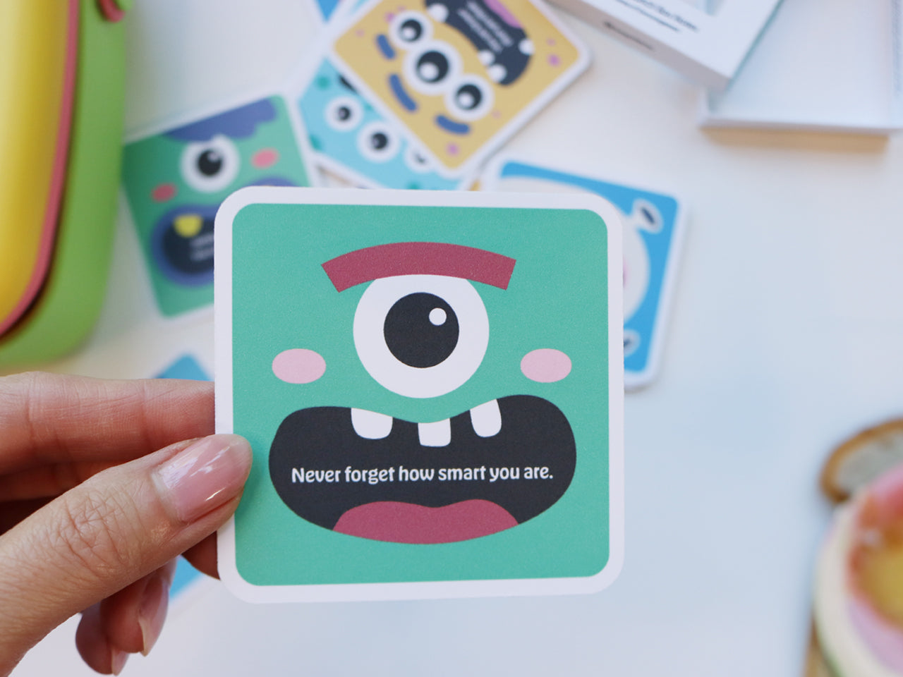 A close-up of a hand holding a green monster-themed lunch box note card with a large cartoon eye and a smile, reading Never forget how smart you are. The background includes scattered monster cards and a green lunchbox.