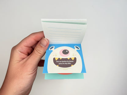 Hand holding an open monster lunch box notes notepad, showing colorful monster artwork on the front cards and lined backs for handwritten messages, handmade kids lunchbox notes for school lunches and backpacks.
