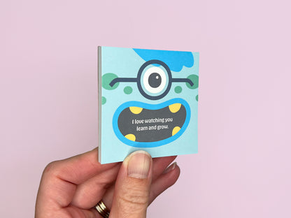 Hand holding a square monster lunch box notes notepad on a soft pink background, showing colorful monster artwork on the front, handmade kids lunchbox notes designed for school lunches and backpacks.