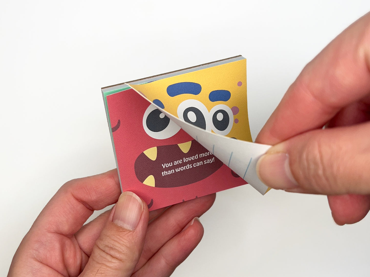 Hands flipping through a square monster lunch box notes notepad, showing colorful monster designs on the front and lined backs for handwritten messages, handmade kids lunchbox notes for school lunches and backpacks.