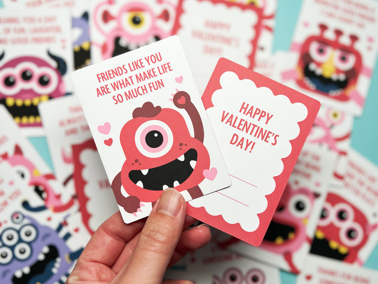 Hand holding two monster-themed Valentine's Day cards in focus, with a colorful array of similar cards blurred in the background. The front of card features a red monster with a single eye and a Valentine message. The back has lines for To and From.