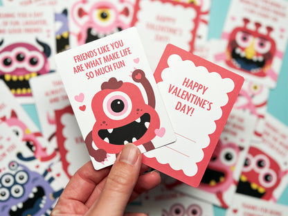 Hand holding two monster-themed Valentine's Day cards in focus, with a colorful array of similar cards blurred in the background. The front of card features a red monster with a single eye and a Valentine message. The back has lines for To and From.