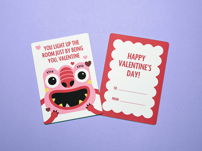 Front and back view of monster-themed Valentine's Day card displayed on a light purple background. Front features a smiling pink monster with expressive eyes, and heart accents. Back has a red scalloped border design and blank spaces for To and From.