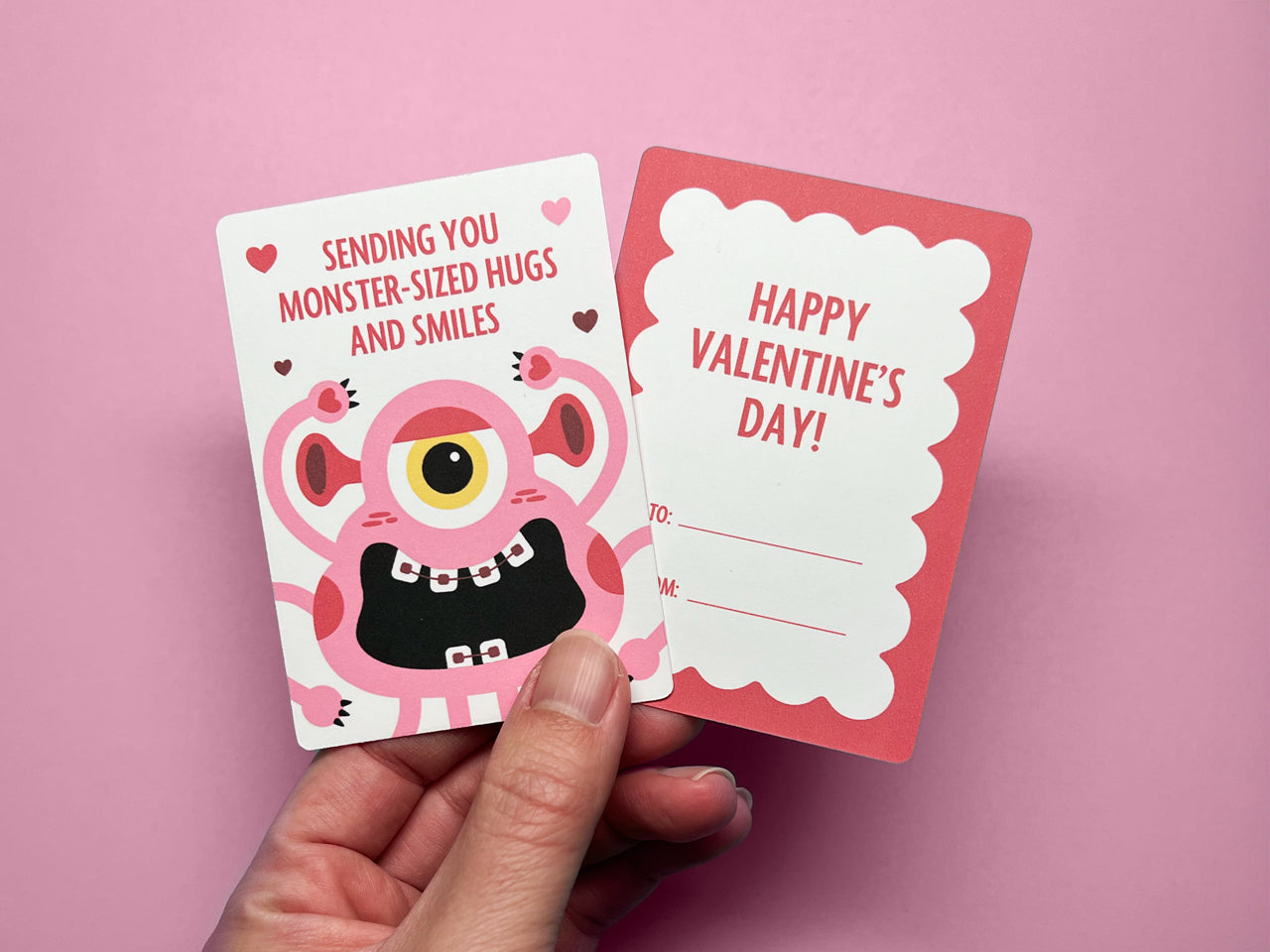 A hand holding monster-themed Valentine's Day cards against a pink background. The front of the card features a cheerful pink monster with one big eye, surrounded by heart accents. The back of card has a scalloped red border with To and From spaces.