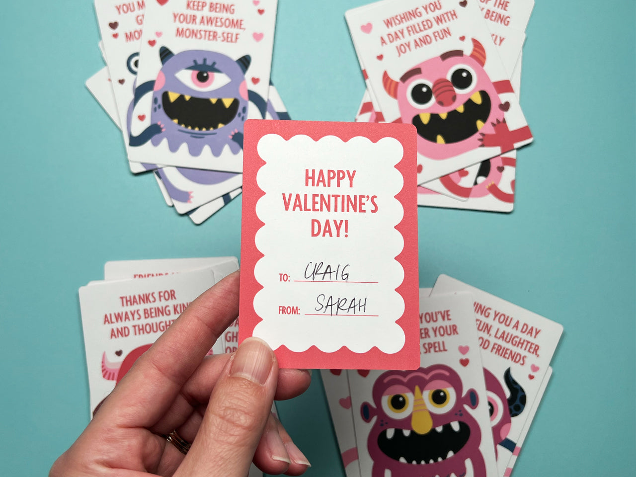 A hand holding the back of a monster-themed Valentine card. The back features a red scalloped border with the text, Happy Valentine's Day! and personalized written in the blank spaces. These cards are perfect for classroom exchanges and parties.