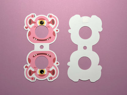 The front and back view of a monster-themed Valentine lollipop holder. The left shows the front design of a pink monster with one eye and a round opening for a lollipop. The right piece shows the backside with adhesive backing and lines for folding.