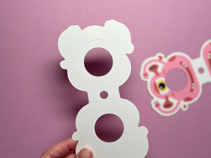 A close-up of the blank backside of a monster-themed Valentine lollipop holder, held in hand against a pink background. The adhesive backing and circular cutouts are visible, with the colorful design of the front blurred in the background.