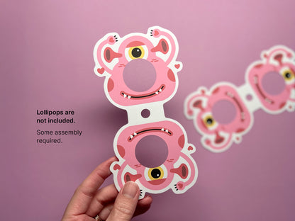 A hand holding a fully printed and colorful monster-themed Valentine lollipop holder with a pink monster design, featuring a round opening for the lollipop. Text on the left reads, Lollipops are not included. Some assembly required.