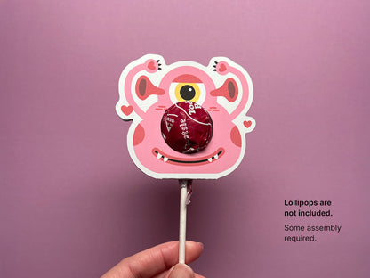 A pink monster-themed Valentine lollipop holder with a red lollipop inserted into the circular opening as a nose. The holder features a smiling monster face with a single eye and heart-shaped details.