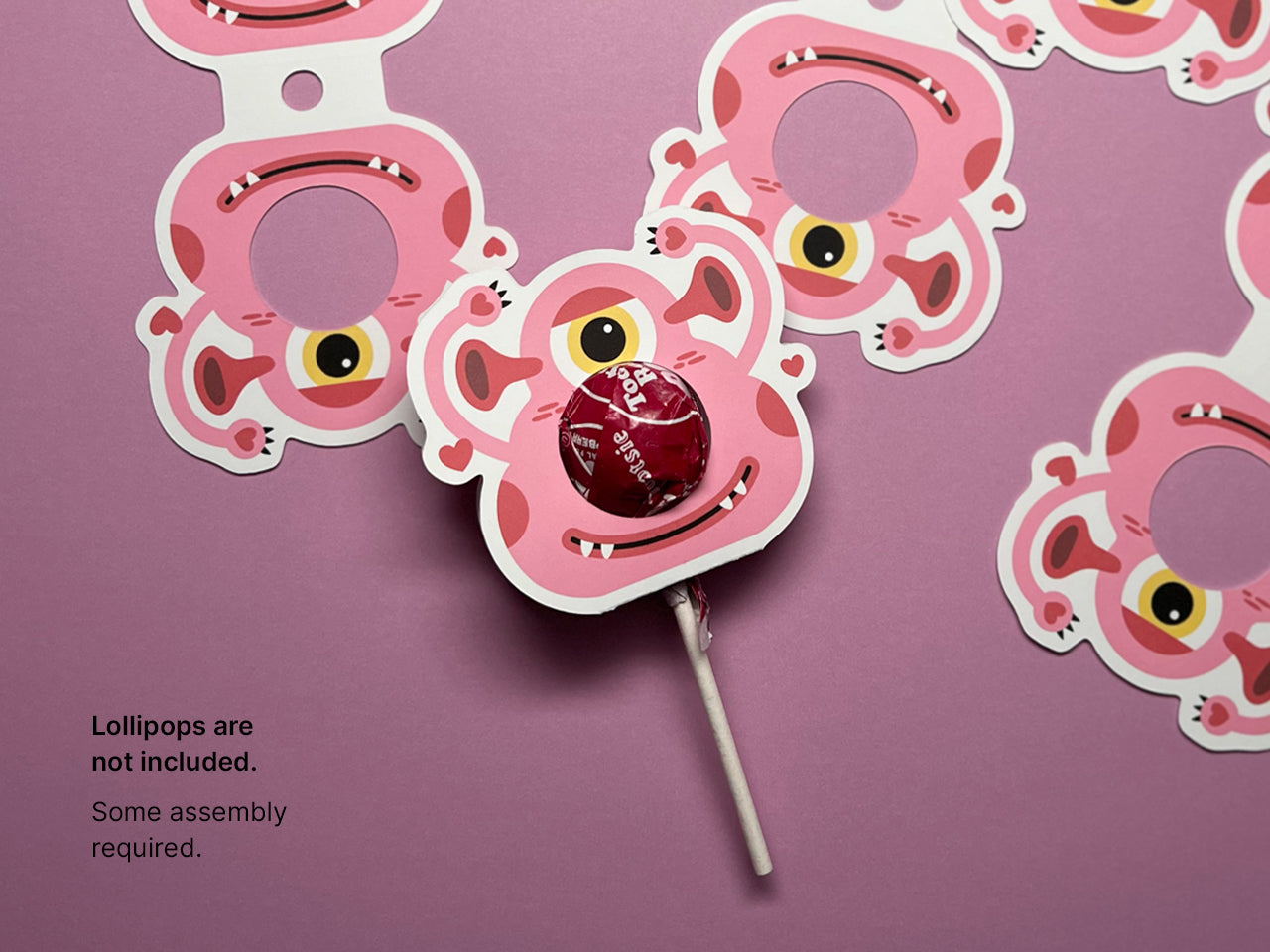 A few pink monster-themed Valentine lollipop holders laid out flat, with one assembled holder featuring a red lollipop displayed in the foreground. The design showcases a playful monster face with a single eye and a smiling expression.