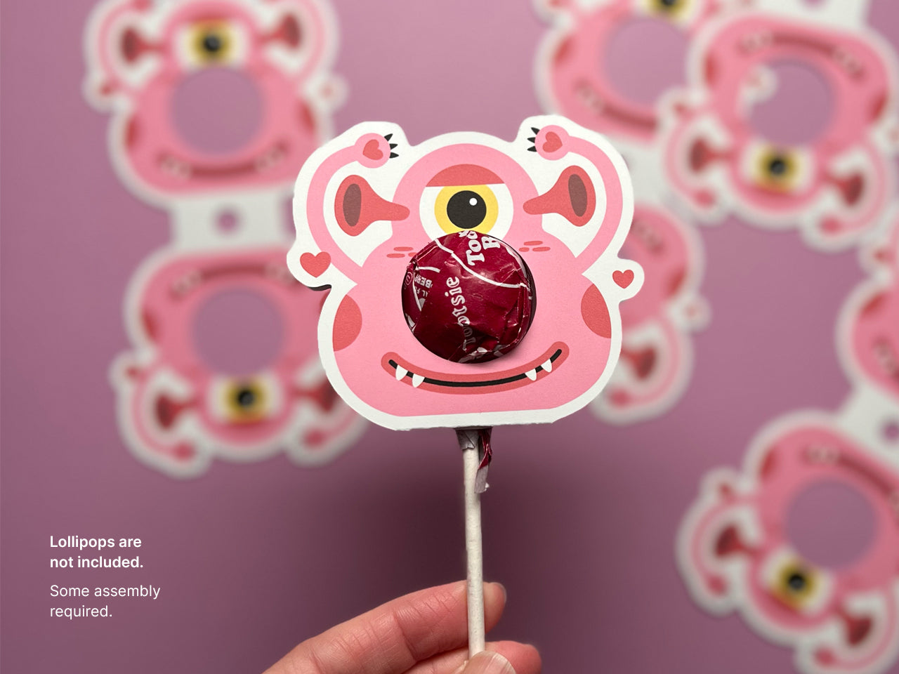 A close-up of a pink monster-themed Valentine lollipop holder with a red lollipop inserted into the circular opening. The playful monster design includes a single eye. Text on the left reads, Lollipops are not included. Some assembly required.