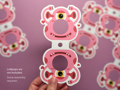 A pink monster-themed Valentine lollipop holder with a circular opening for a round lollipop. The design features a cheerful one-eyed monster with heart accents. Text on the left reads, Lollipops are not included. Some assembly required.