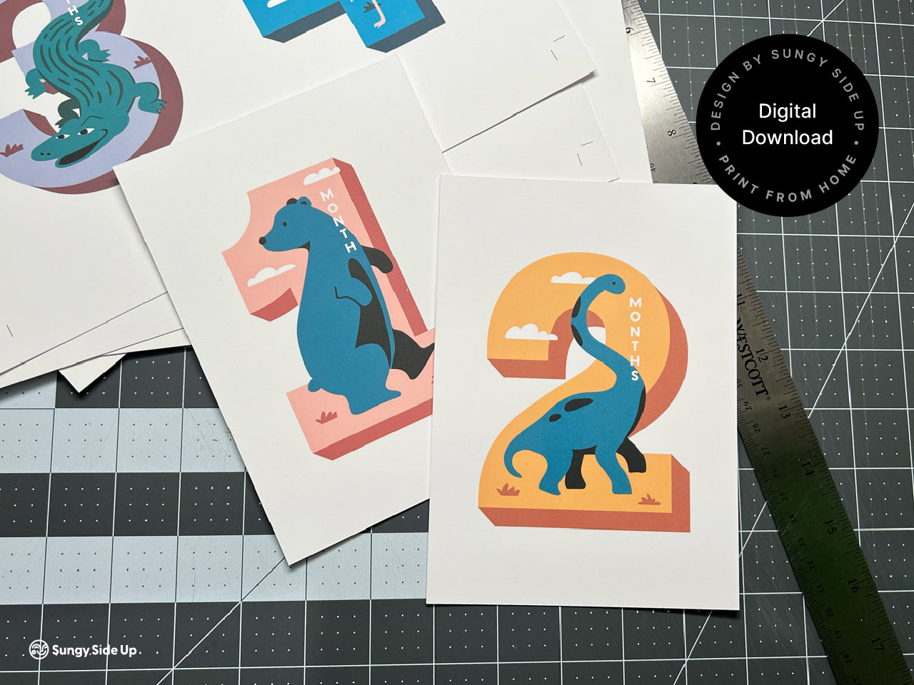 A set of printable monthly milestone cards featuring vibrant, animal-themed designs for each month from 1 to 12. Cards are arranged on a cutting mat, showcasing colorful illustrations like a dinosaur, flamingo, and alligator.