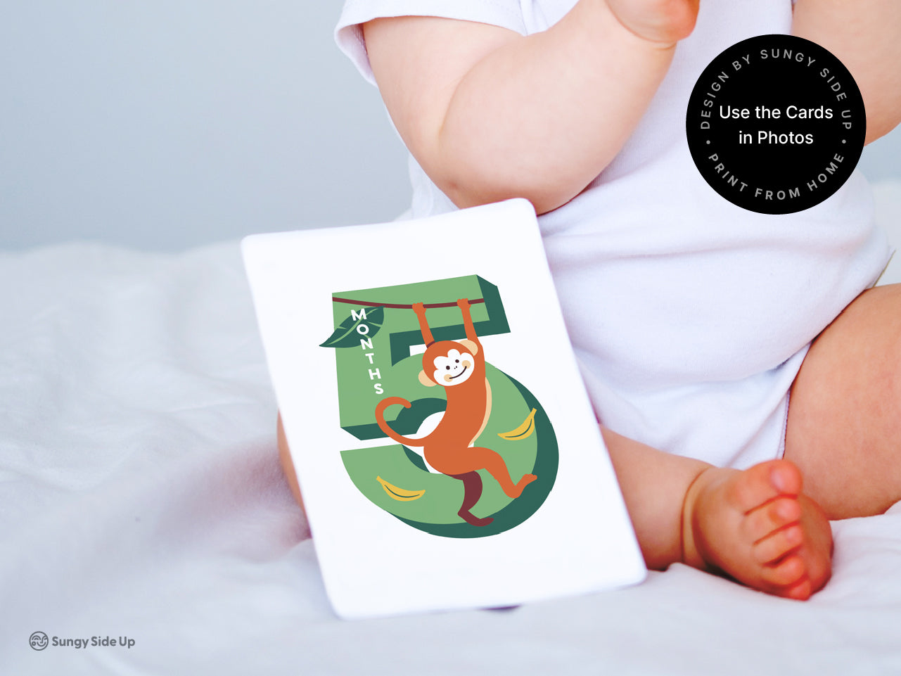 A baby in a white one piece with a printable milestone card for 5 months with a cheerful monkey illustration. The monthly milestone card is printed at home on cardstock and styled as part of a photo to showcase its use for baby milestone photography.