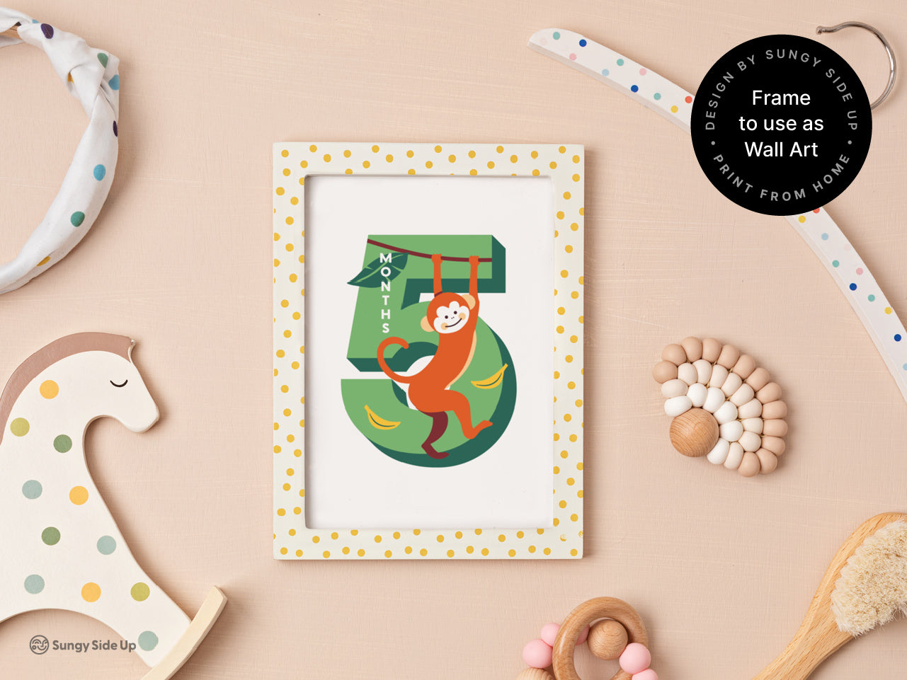 A framed printable monthly milestone card for 5 months, featuring a monkey design and bananas. Displayed on a pastel pink background with playful nursery decor, including a wooden rocking horse and baby toys, highlighting its dual use as wall art.