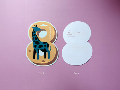 A giraffe-themed die-cut milestone card in the shape of the number 8. The front features a playful illustration of a giraffe, while the back includes prompts to document baby details like height, weight, favorite food, and milestones.