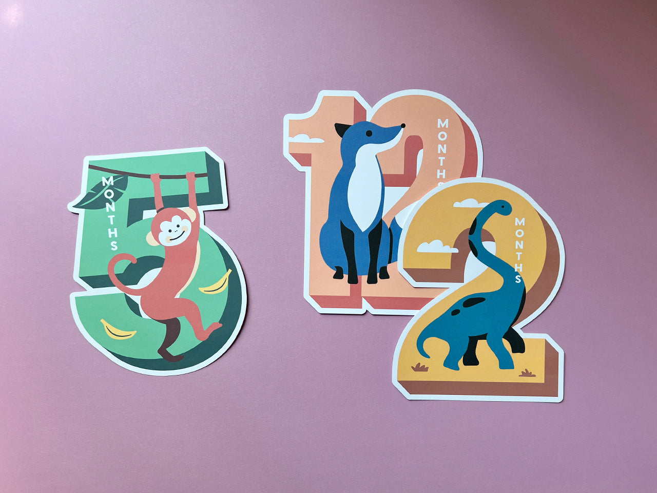 A collection of colorful die-cut milestone cards featuring playful animals: a monkey for 5 months (green background with bananas), a fox for 12 months (peach background), and a dinosaur for 2 months (golden background).