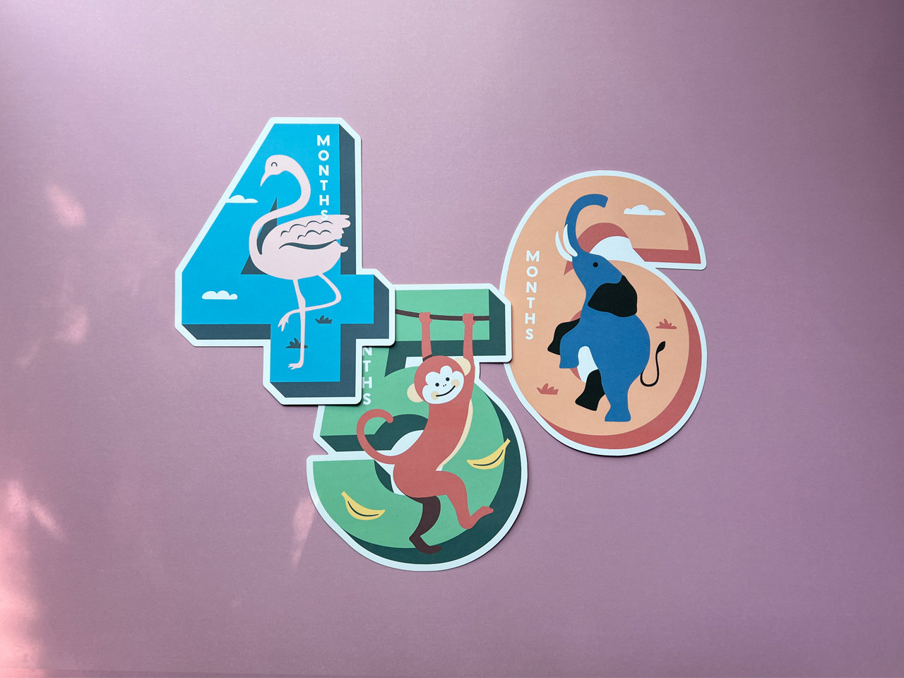 Three colorful die-cut milestone cards featuring animal illustrations: a flamingo for 4 months (blue background), a monkey for 5 months (green background with bananas), and an elephant for 6 months (peach and red background). Ideal for monthly baby photos and keepsake memory collections.