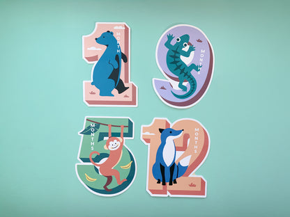 A collection of four colorful die-cut milestone cards featuring animal illustrations: a bear for 1 month, a crocodile for 9 months (purple background), a monkey for 5 months (green background with bananas), and a fox for 12 months (peach background).