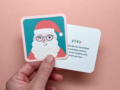 A person holding a square mini note with a Santa Claus illustration on the front, smiling and winking. Behind the Santa card, another note is partially visible with holiday lights and text reading, "It’s a cozy day for storytelling! Share a holiday memory or start a new tradition with someone you love."