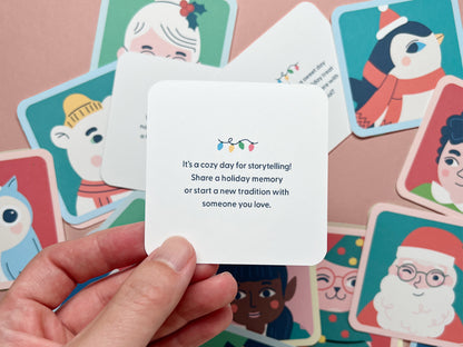 A hand holding a square mini note with text that says, "It’s a cozy day for storytelling! Share a holiday memory or start a new tradition with someone you love," accompanied by a string of holiday lights. In the background, several other mini notes are spread out, each featuring unique holiday characters like elves, animals, and other festive designs.