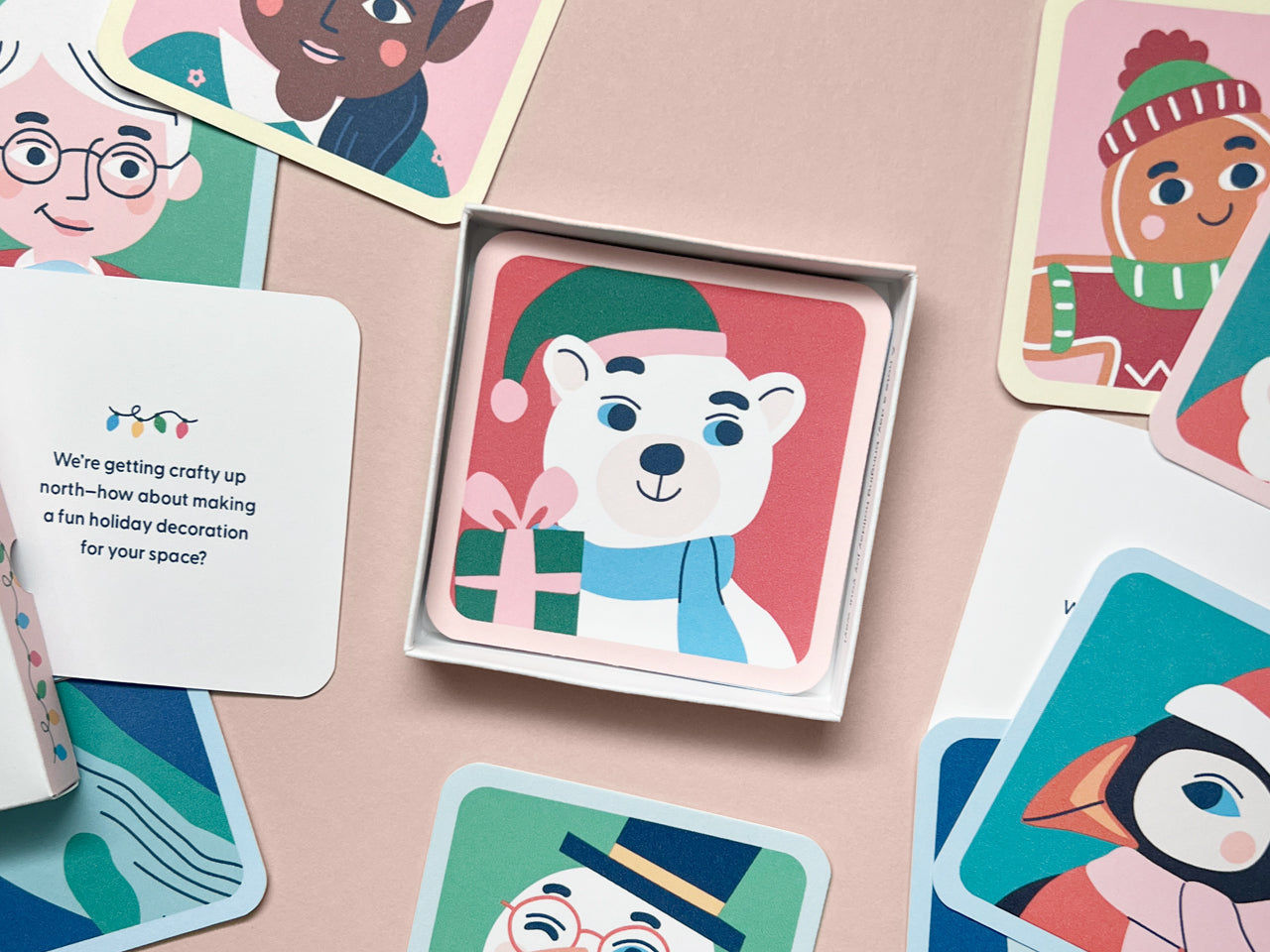 A square mini note featuring a polar bear wearing a green hat and holding a gift, neatly placed in a small gift box. Surrounding the box are various mini notes with other character illustrations and holiday-themed text, spread out on a pastel pink background.