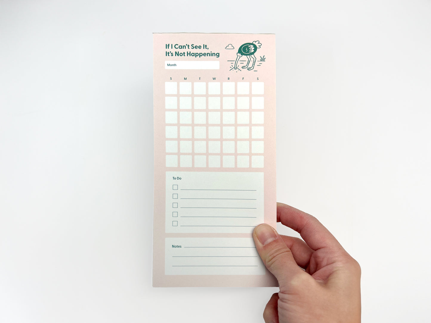 Front-facing photo of a tall, narrow monthly planner notepad held upright in one hand against a white background. The notepad has a soft pink background with the phrase “If I Can’t See It, It’s Not Happening” at the top and a small illustrated ostrich with its head in the ground. Below the title is a blank month label, a full monthly calendar grid with weekday initials, a lined to-do list with checkboxes, and a lined notes section at the bottom.