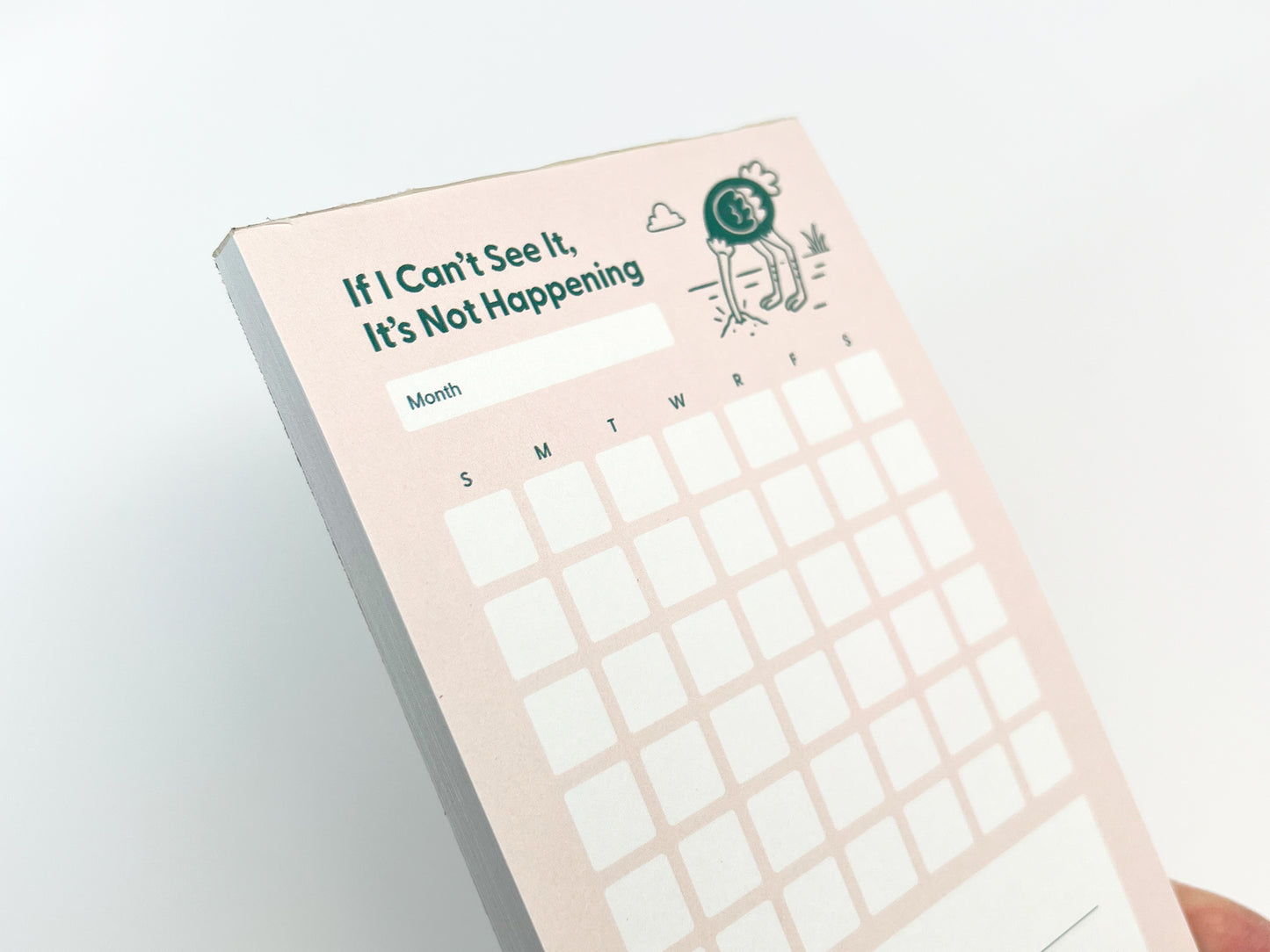Angled close-up of the top portion of a tall monthly planner notepad with a soft pink background. The notepad reads “If I Can’t See It, It’s Not Happening” above a blank month label, with a small illustrated ostrich burying its head in the ground near the title. The top rows of the monthly calendar grid are visible, and the stacked paper edge is shown along the left side against a white background.